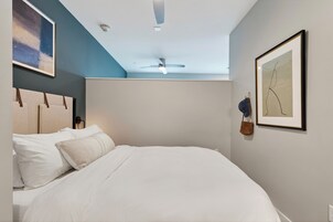 A bedroom with a bed, a ceiling fan, a framed abstract painting, and a hanging bag.