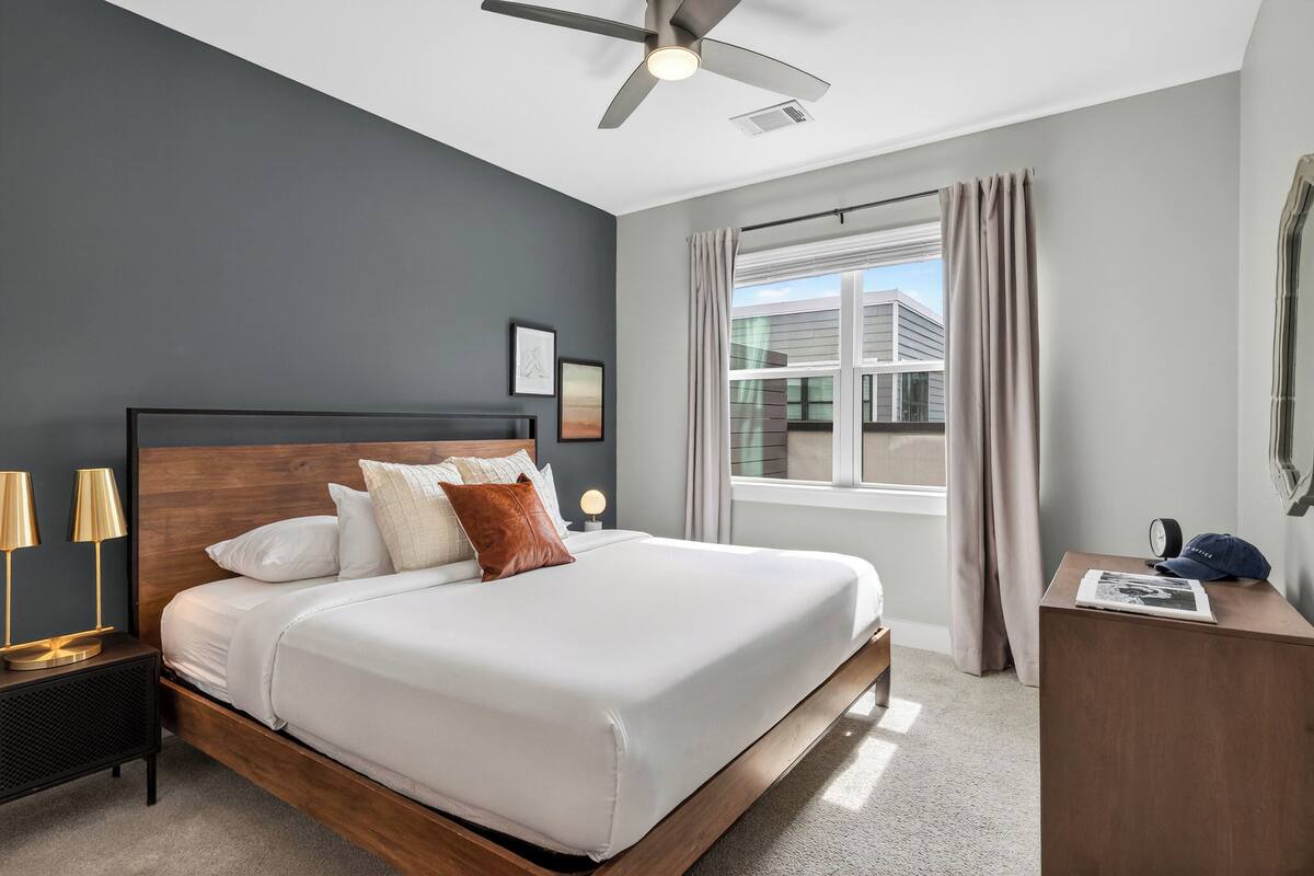 A bedroom with a bed, bedside lamps, a nightstand, a ceiling fan, a window with curtains, and a view of buildings outside.