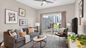 A modern living room with a grey sofa, a coffee table, and a TV. There is a balcony with a view of buildings.