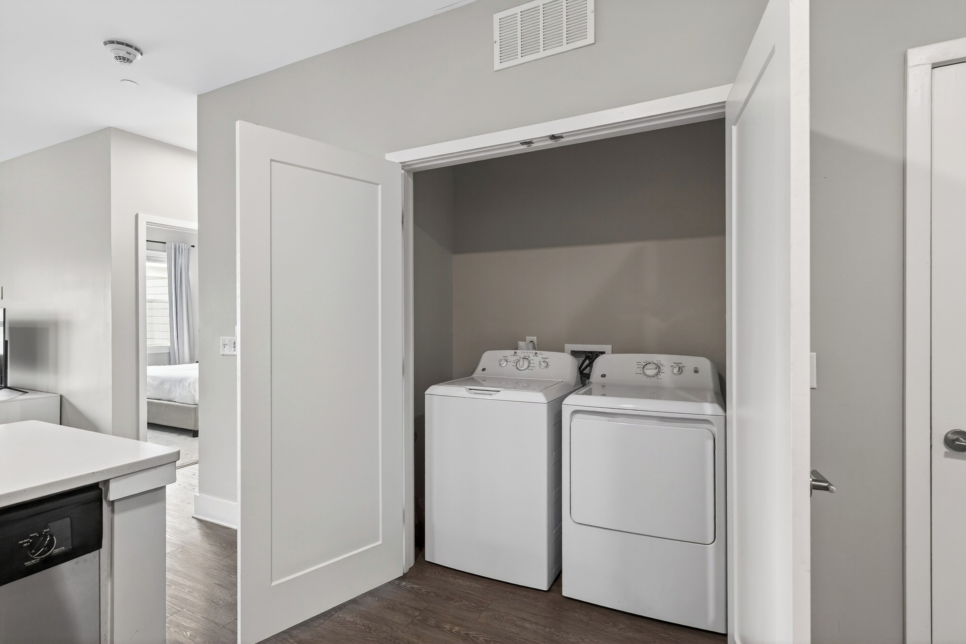A laundry room with two white washing machines and a dryer, a white countertop with a dishwasher, and a door leading to a bedroom.