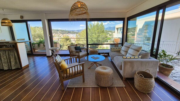 Un salon moderne comprenant un canapé, des fauteuils, une petite table et un luminaire suspendu. De grandes fenêtres offrent une vue sur la ville et les montagnes.