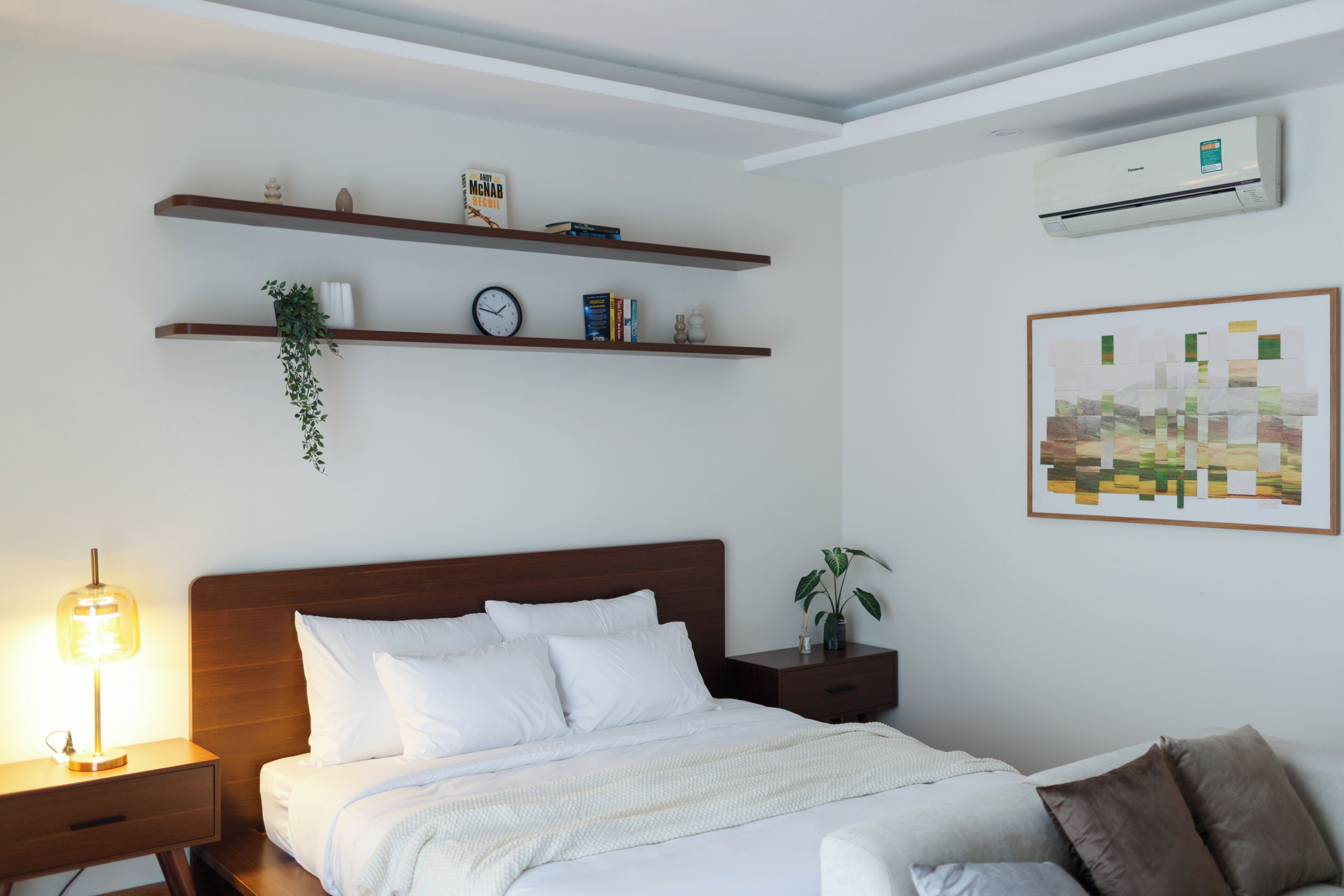 Un dormitorio con cama, mesitas de noche, aire acondicionado instalado en la pared y estantes con libros y objetos decorativos.
