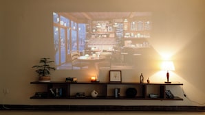 A room with a projection of a café interior, a shelf with books and decorative items, a potted plant, and a lamp. - Beloved Summer - 4BR Cozy & Peaceful with Elevator (Hà Nội)