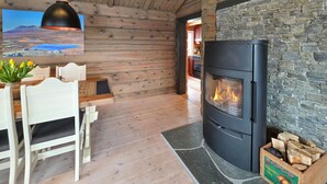 A cozy cabin interior with a stone wall, a wood-burning stove, a dining area with a table and chairs, and a painting of a landscape. - Amazing home in Ustaoset (Ustaoset)