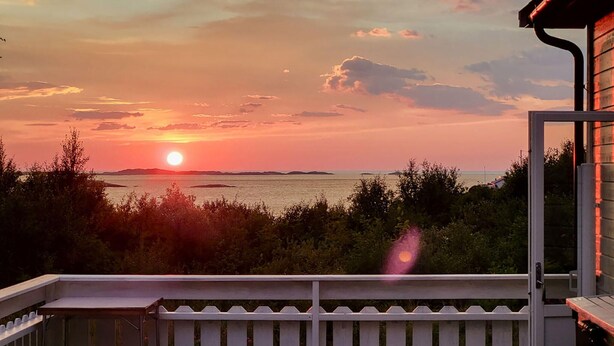 Un balcone con vista sul tramonto sul mare, una recinzione bianca e un edificio con tetto rosso.