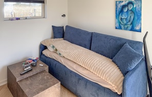 Un salon avec un canapé bleu, un plaid beige, une petite table avec des télécommandes et un tableau accroché au mur.
