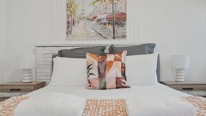 Una cama impecablemente hecha con un plaid estampado, un cuadro de paisaje urbano y una chimenea de ladrillo blanco.