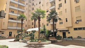 Un immeuble résidentiel à plusieurs étages doté de balcons, d’un espace extérieur couvert avec des sièges et d’une cour pavée agrémentée de palmiers et de bancs.