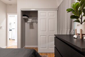 Ein Schlafzimmer mit einem Bett, einem Schminktisch, einem Kleiderschrank mit Kleidung und einer Zimmerpflanze.