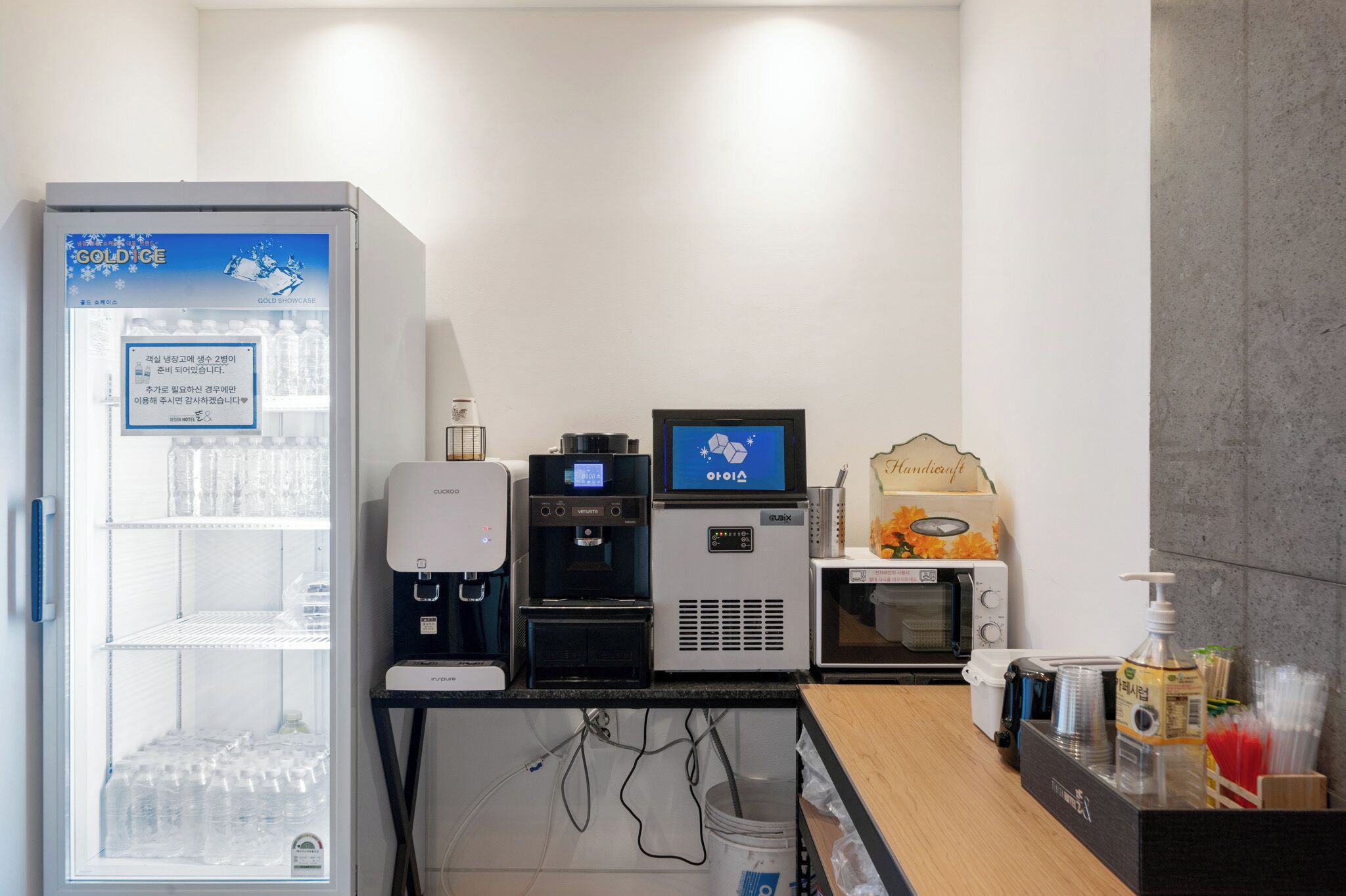 A hotel reception area with a refrigerator, a coffee machine, a water dispenser, a digital screen, a microwave, and a small display case.