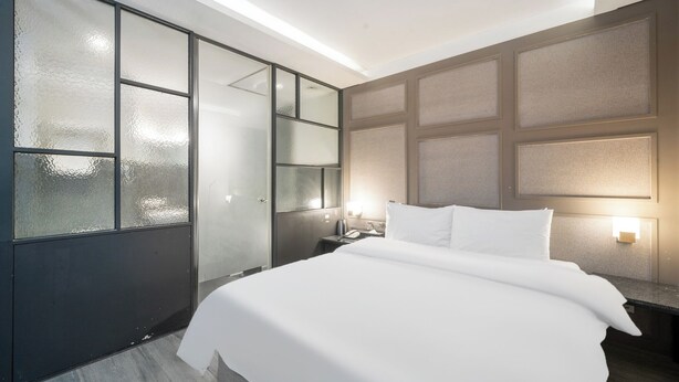 A modern hotel room with a large bed, a nightstand, a wall-mounted lamp, and a frosted glass partition. - Design Hotel Tteulaen (Pyeongtaek)