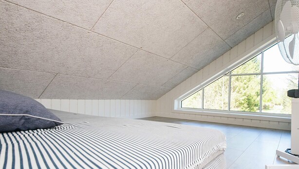 Une chambre avec un plafond en pente, un lit avec une literie à rayures, une fenêtre donnant sur des arbres et un ventilateur sur pied.