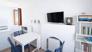 Ein Zimmer mit weißem Schreibtisch, Stühlen, einem Fernseher, einer Heizung und einem Bücherregal voller Bücher.