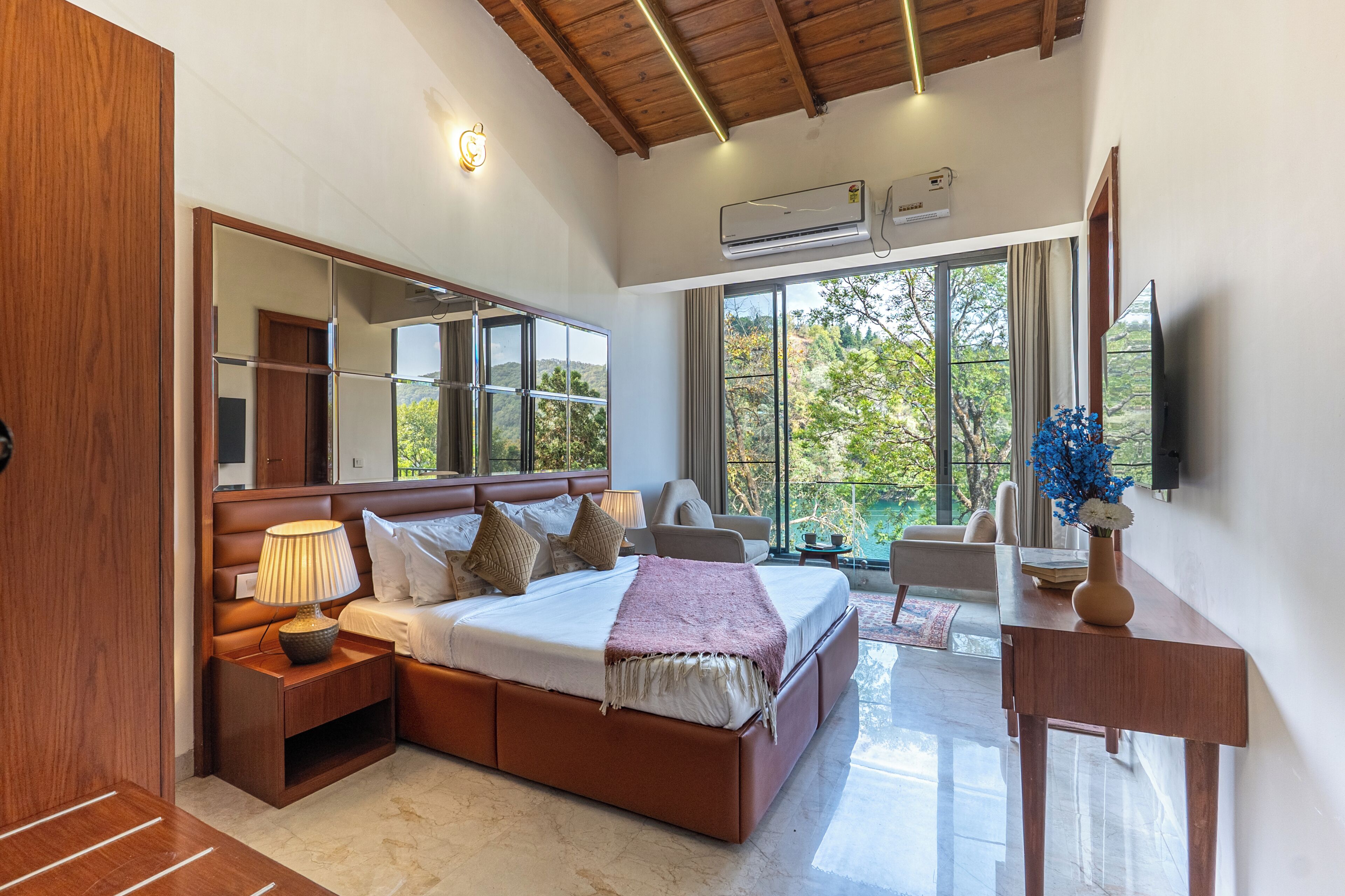 A bedroom with a large bed, bedside tables, a mirror, a TV, and a window with a view of trees.