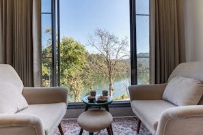 A room with two sofas facing a large window with a view of trees and hills. - ALAYA STAYS Lakefront Suite Naukuchiatal (Siloti Pant)