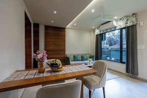 A modern dining area with a wooden table, chairs, a vase with flowers, and a view of the outdoors through a large window. - ALAYA STAYS Lakefront Suite Naukuchiatal (Siloti Pant)