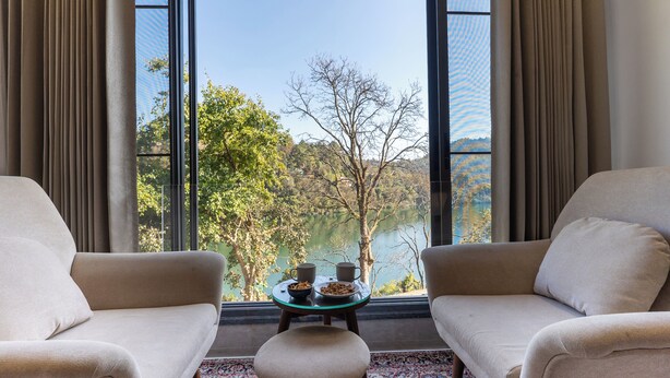 Habitación con dos sofás frente a una mesa con un bol y dos tazas, y vistas a un lago y árboles a través de amplias ventanas.