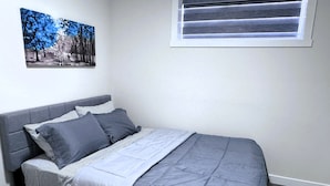 A bedroom with a bed, a window with blinds, and a painting on the wall. - The Suite Spot (Airdrie)