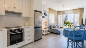 A modern kitchen with white cabinetry, a built-in oven, and a refrigerator. There is a dining area with a blue table and chairs, and a balcony with a view of the sea. - Seawaves 1- BR Apt. by Meneou Beach (Meneou)
