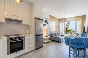 A modern kitchen with white cabinetry, a built-in oven, and a refrigerator. There is a dining area with a blue table and chairs, and a balcony with a view of the sea. - Seawaves 1- BR Apt. by Meneou Beach (Meneou)