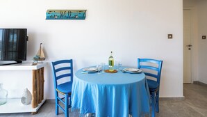 A dining area with a round table, blue chairs, a television on a white cabinet, and a wall-mounted artwork. - Seawaves 1- BR Apt. by Meneou Beach (Meneou)