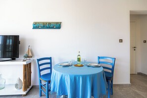 A dining area with a round table, blue chairs, a television on a white cabinet, and a wall-mounted artwork. - Seawaves 1- BR Apt. by Meneou Beach (Meneou)