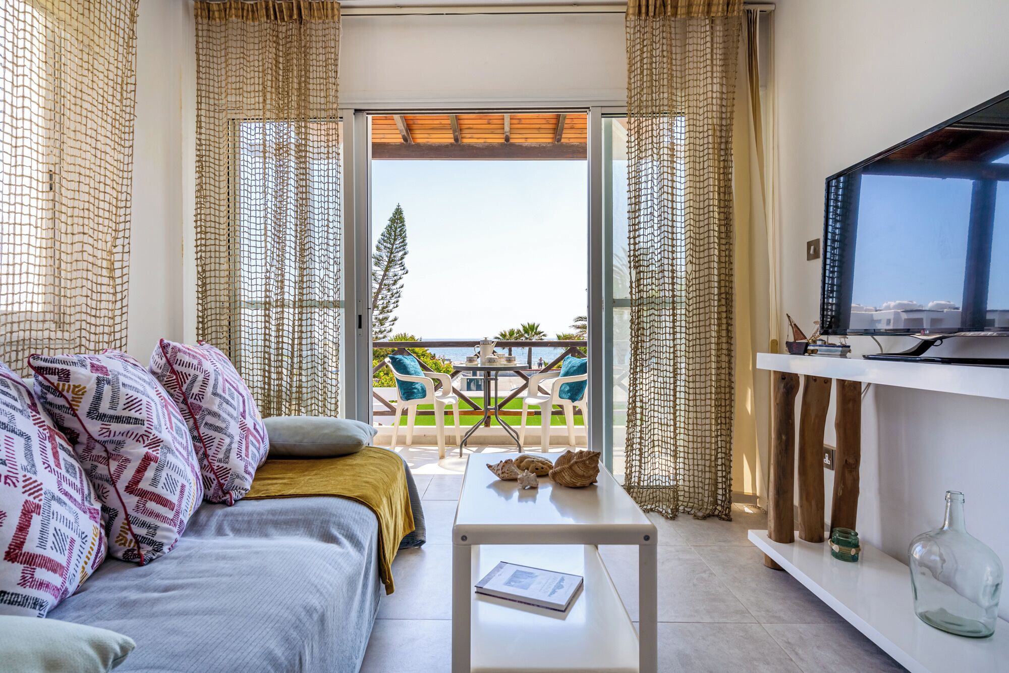 A modern living room with a sofa, a coffee table, and a TV. There is a balcony with a table and chairs.