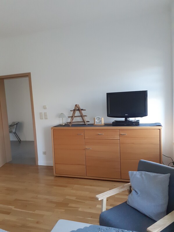Ein Zimmer mit einem Holzkasten, einem Fernseher darauf, einem Stuhl mit Kissen und einer Tür zu einem weiteren Raum.
