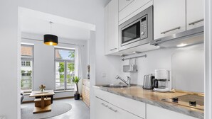 Une cuisine moderne avec des meubles blancs, un four à micro-ondes encastré, une machine à café et une vue sur l’extérieur par une fenêtre.