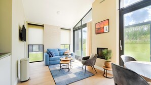 Ein modernes Wohnzimmer mit einem blauen Sofa, einem grauen Sessel, einem kleinen runden Tisch und einem großen Fenster mit Blick ins Freie.