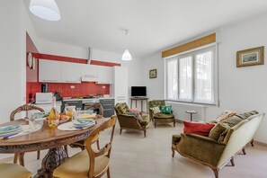 Eine moderne Küche mit weißen Schränken, einem Esstisch gedeckt mit Tellern, Gläsern und einer Flasche, sowie ein Wohnbereich mit Sofa und Sesseln.