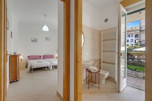 Une chambre à coucher avec un lit, des tables de chevet, un luminaire suspendu, une salle de bain avec un lavabo et des toilettes, et une vue sur les bâtiments à travers une porte ouverte.