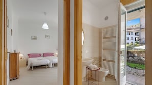 A bedroom with a bed, bedside tables, a hanging light fixture, a bathroom with a sink and toilet, and a view of buildings through an open door. - Casa Adelina, Center Flat With Private Garden (Stresa)