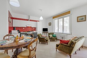 Une cuisine moderne avec des meubles blancs, une table à manger dressée avec des assiettes, des verres et une bouteille, et un coin salon avec un canapé et des fauteuils.