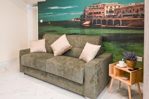 A living room with a sofa, a small wooden side table with a cup and a remote control, and a large wall mural of a cityscape. - Walk to Balluta & Spinola - Modern 2bdr Apart (St. Julian's)