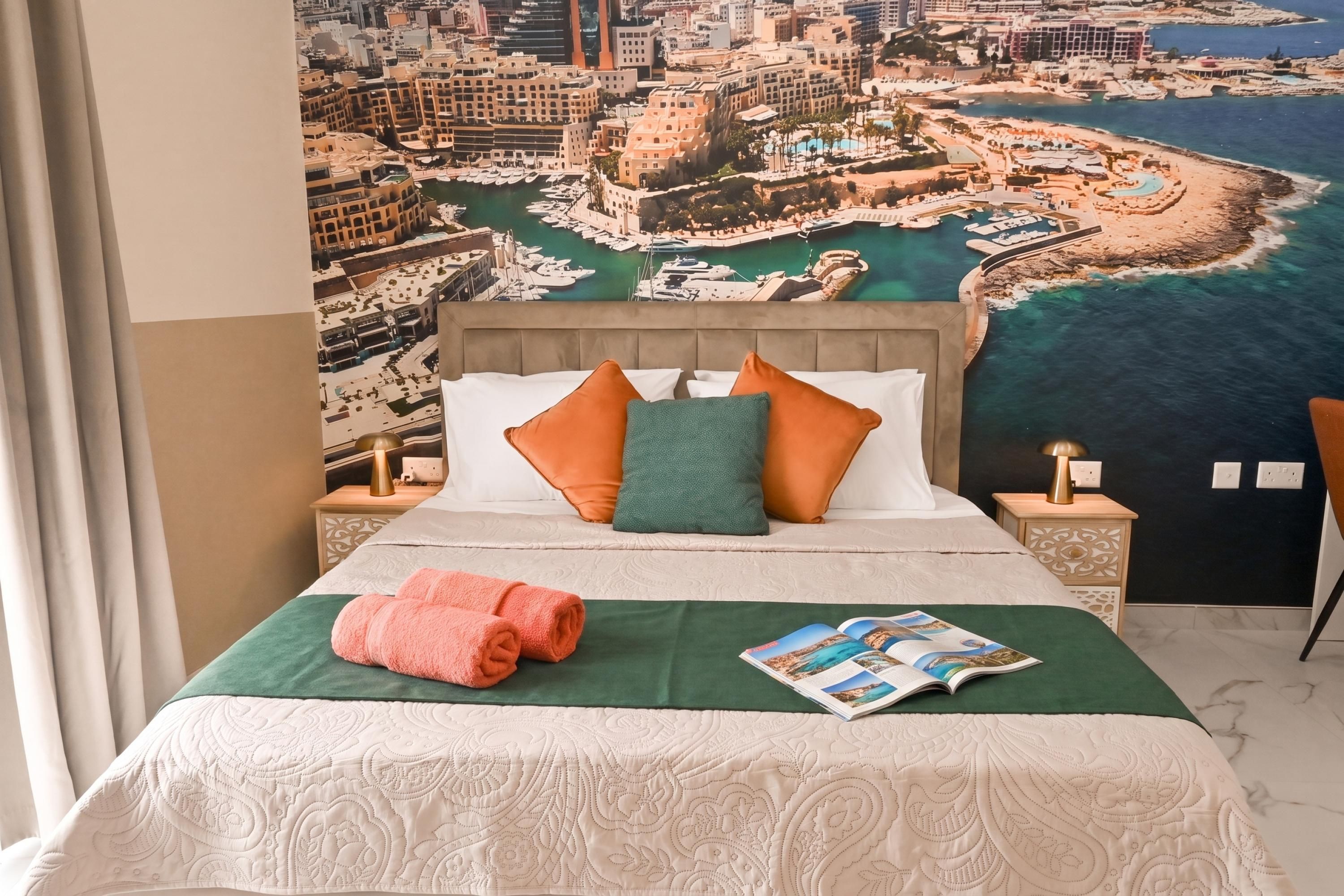 A hotel room with a bed, pillows, a green bedspread, a magazine, and a view of a coastal cityscape.