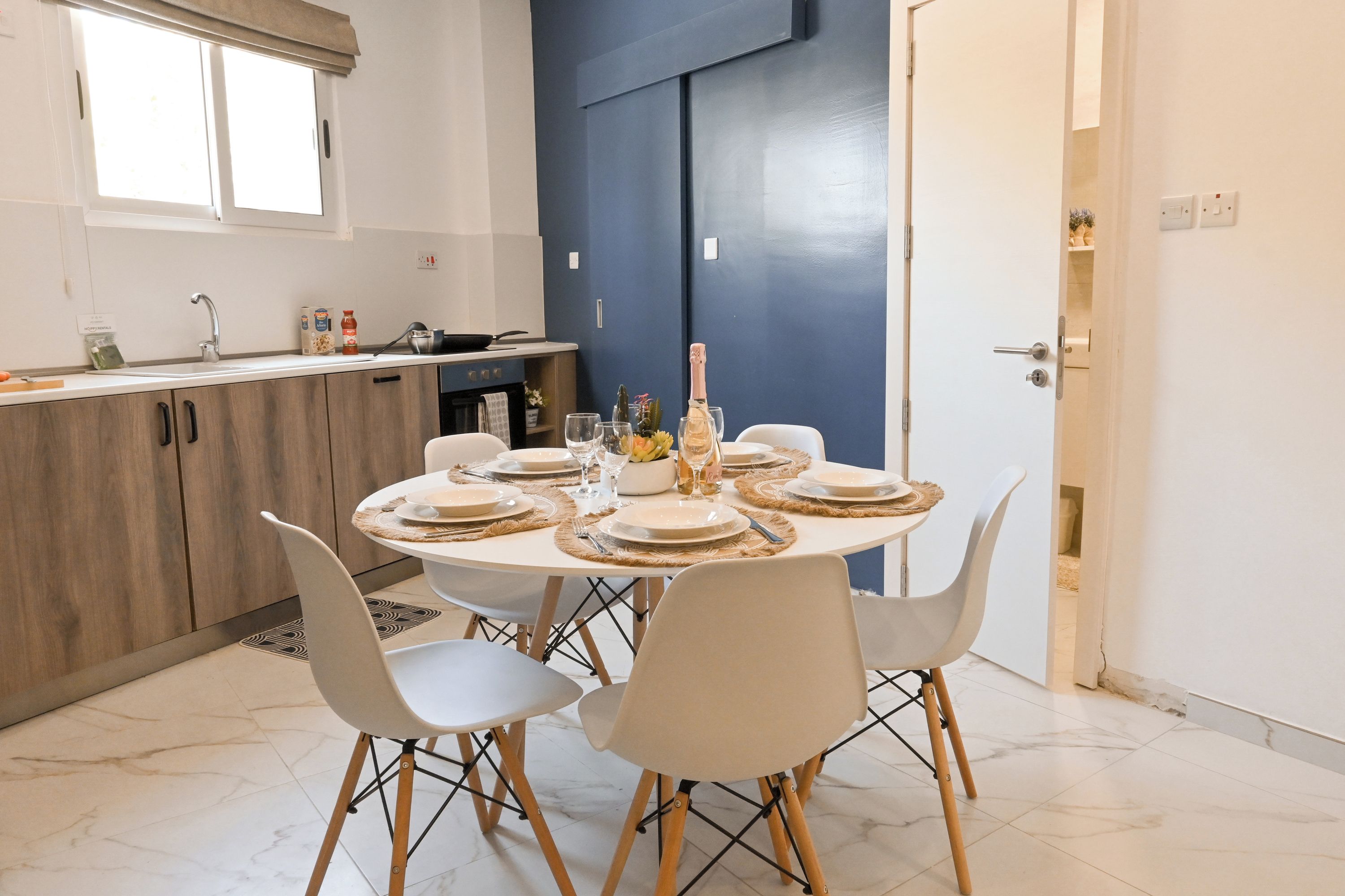 A dining area with a round table set for four, white chairs, a kitchen in the background, and a blue door.
