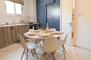 A dining area with a round table set for four, white chairs, a kitchen in the background, and a blue door. - Walk to Balluta & Spinola - Modern 2bdr Apart (St. Julian's)