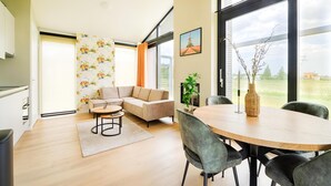 Un salon moderne comprenant un canapé d’angle, une table à manger avec des chaises, un coin cuisine et de grandes fenêtres donnant sur l’extérieur.