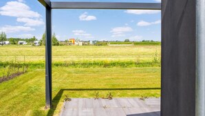 Vue depuis une pièce dont le sol est en béton et qui présente des poutres métalliques, donnant sur un champ verdoyant avec quelques bâtiments visibles au loin.