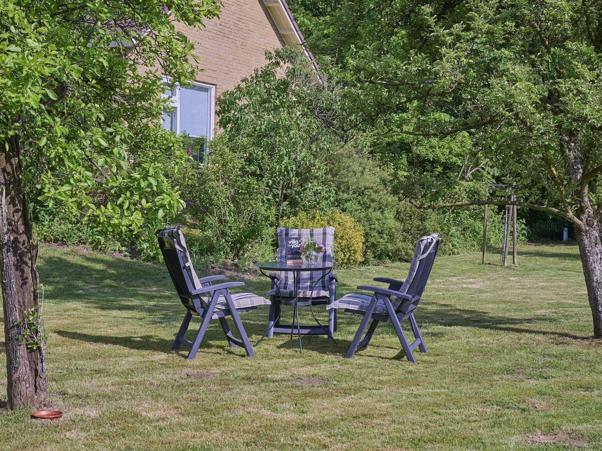 An outdoor seating area with a table and four chairs, surrounded by trees and grass.
