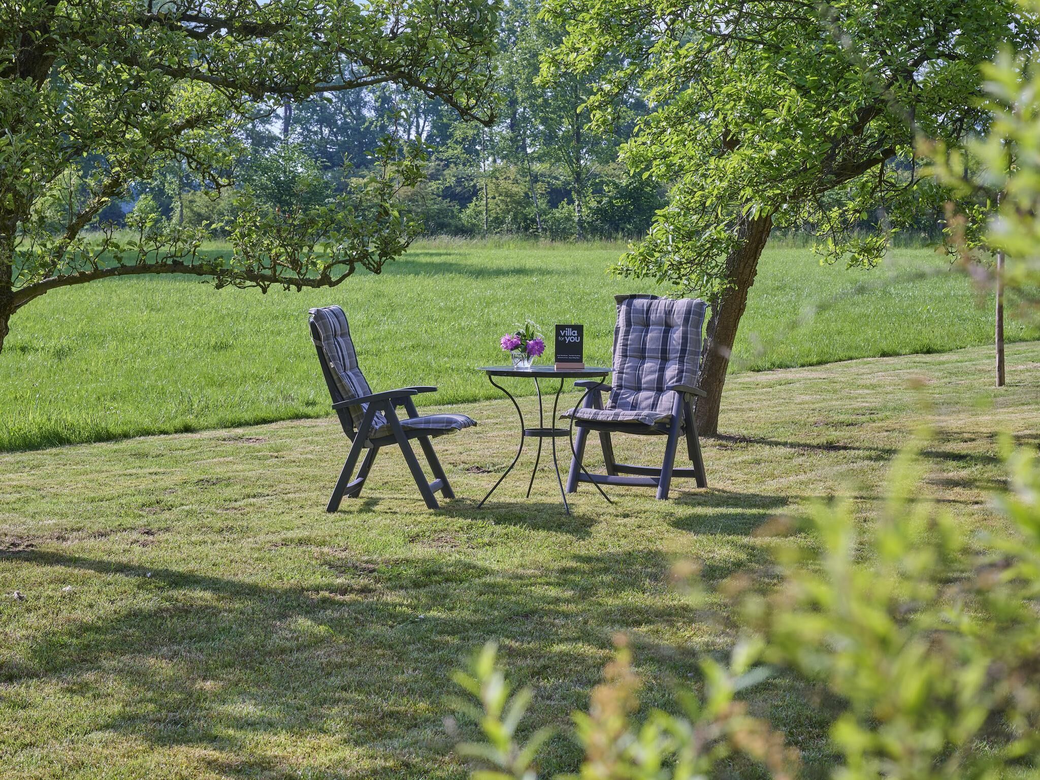 An outdoor seating area with two chairs and a table, surrounded by lush greenery and trees.