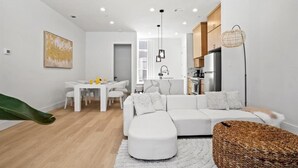 Un salon moderne avec un canapé blanc, une table à manger avec des chaises, un coin cuisine avec des placards et un luminaire suspendu.
