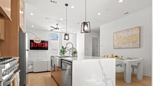 Une cuisine moderne dotée d’un îlot central, d’un coin repas avec une table et des chaises blanches, d’un ventilateur de plafond et d’une œuvre d’art accrochée au mur.