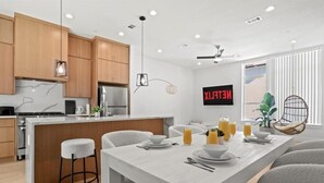 Une cuisine moderne avec des meubles en bois, un îlot central doté de tabourets de bar, un coin repas avec une table blanche dressée, et un téléviseur fixé au mur qui affiche « NETFLIX ».