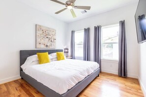 Une chambre à coucher comprenant un lit, un ventilateur de plafond, une fenêtre avec des rideaux et une œuvre d’art accrochée au mur.