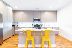 Una cocina moderna con gabinetes grises, una isla central, dos taburetes amarillos y un refrigerador de acero inoxidable.