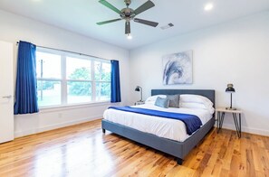 Un dormitorio moderno con una cama grande, ventilador de techo, ventana con cortinas y una obra de arte en la pared.