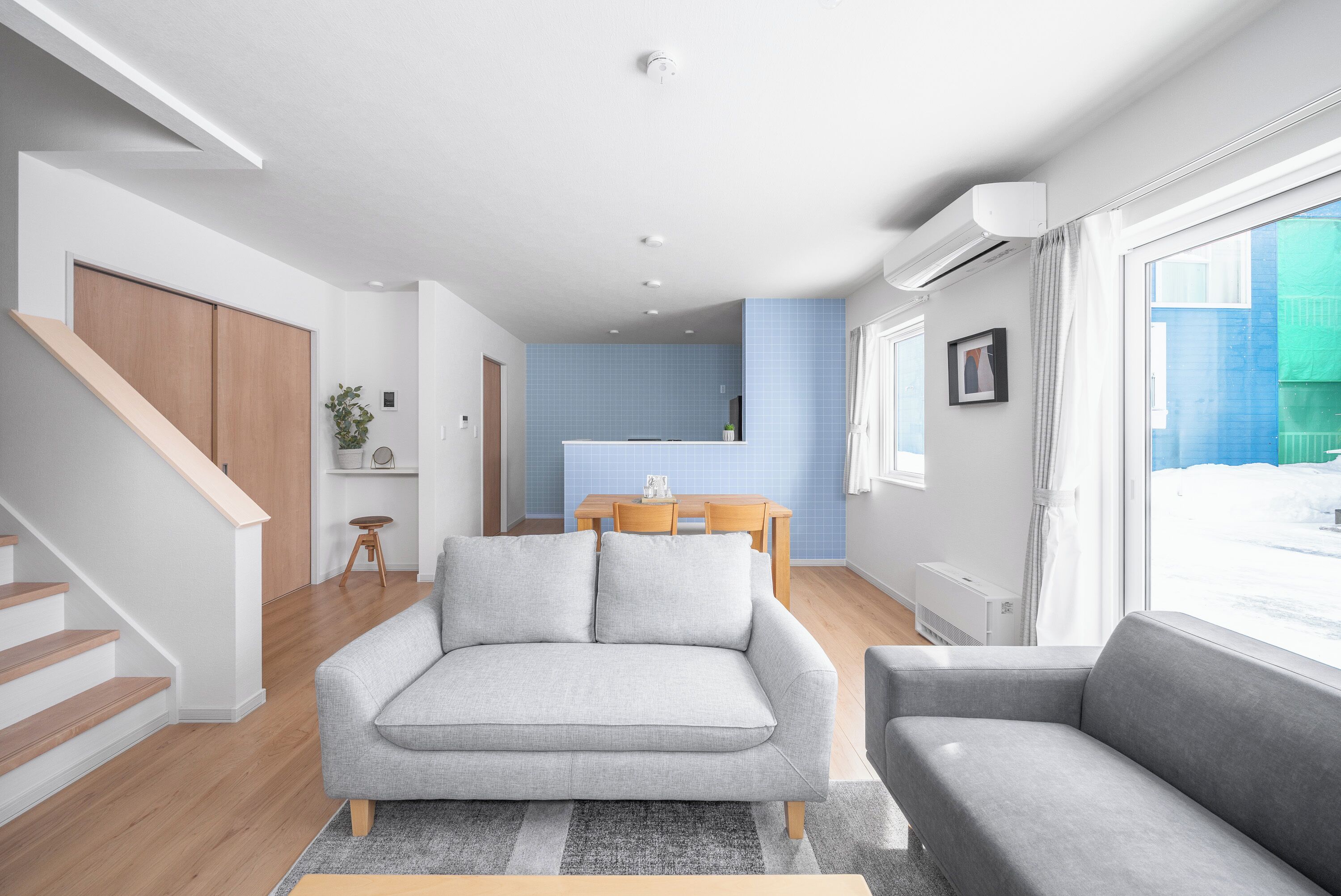 A modern living room with a grey sofa, a wooden staircase, a dining area with a blue tiled wall, and a large window.