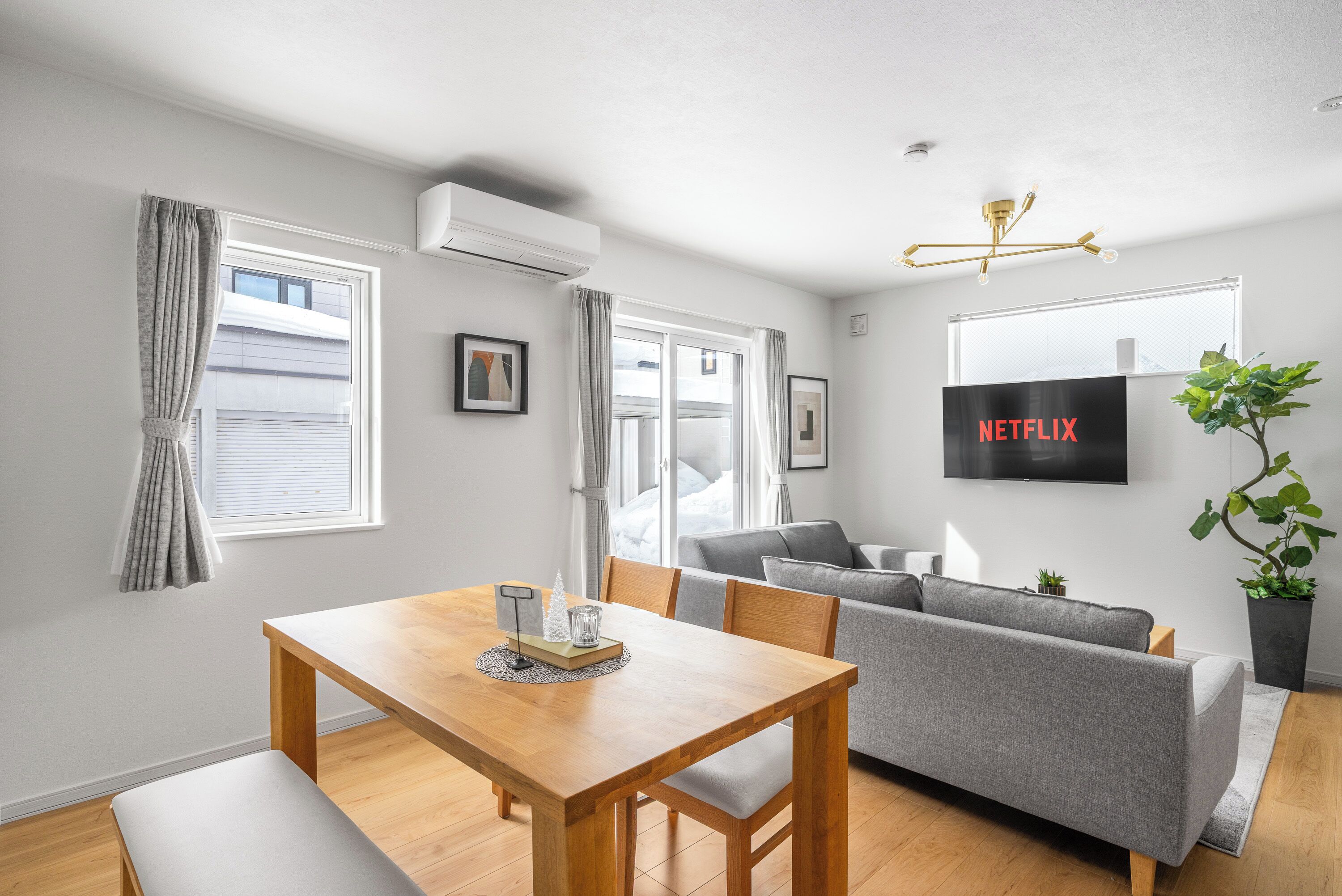 A modern living room with a wooden dining table, grey sofa, and a mounted TV displaying 'NETFLIX'.